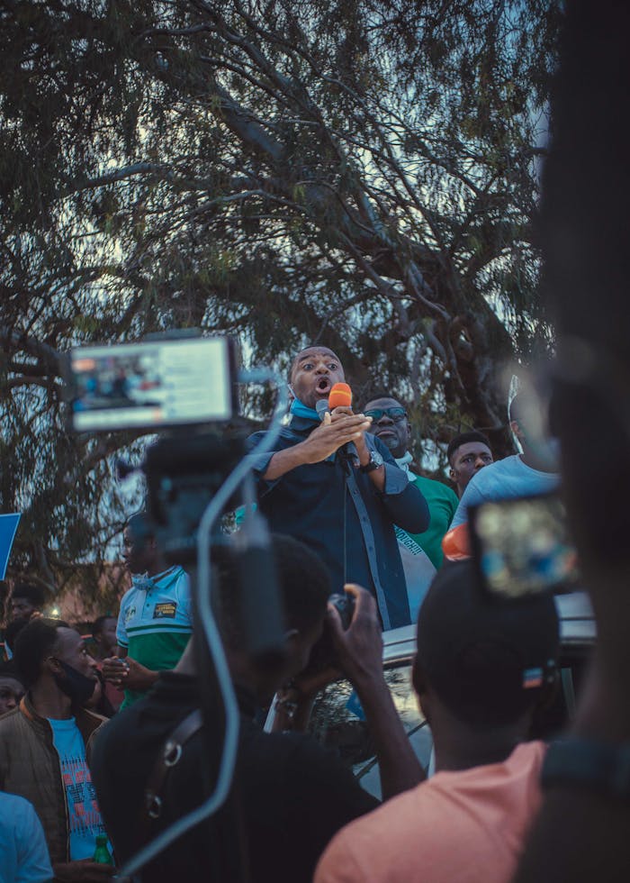 A speaker addresses a crowd with passion, captured through various recording devices outdoors.